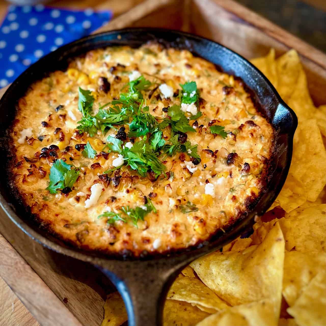 Black cast iron skillet with Elote Dip sitting in a wooden tray surrounded by tortilla chips with blue napkins in the background. 