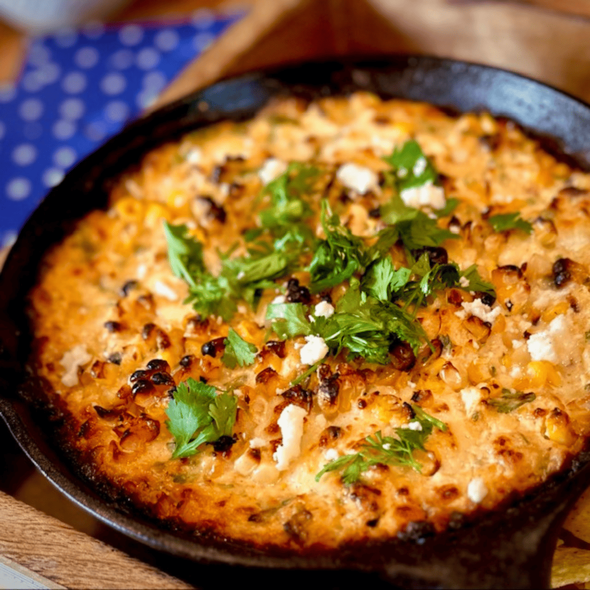 Black cast iron skillet with elote dip sitting in a wooden tray with blue napkins in the background.