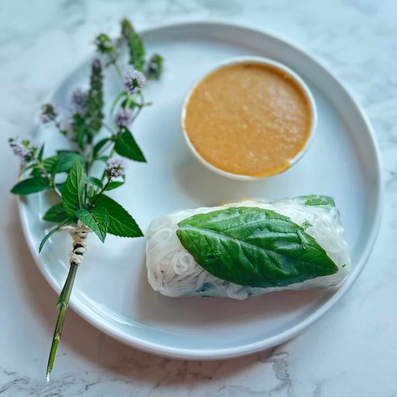 Fresh roll with a Thai Basil leaf sitting on a white plate next to a ramekin of peanut sauce and a sprig of fresh mint.