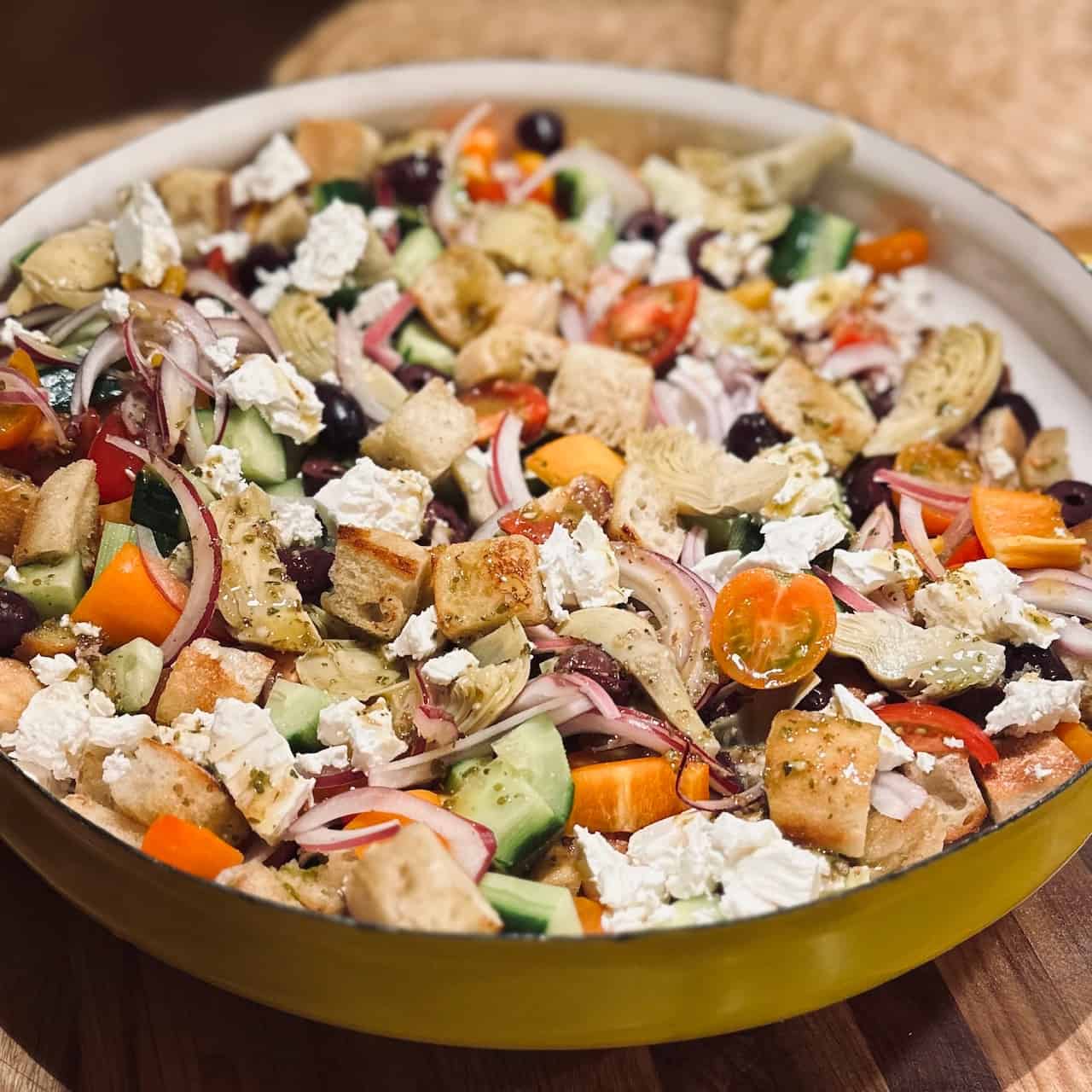 Greek Panzanella in a yellow dish on a wooden cutting board.