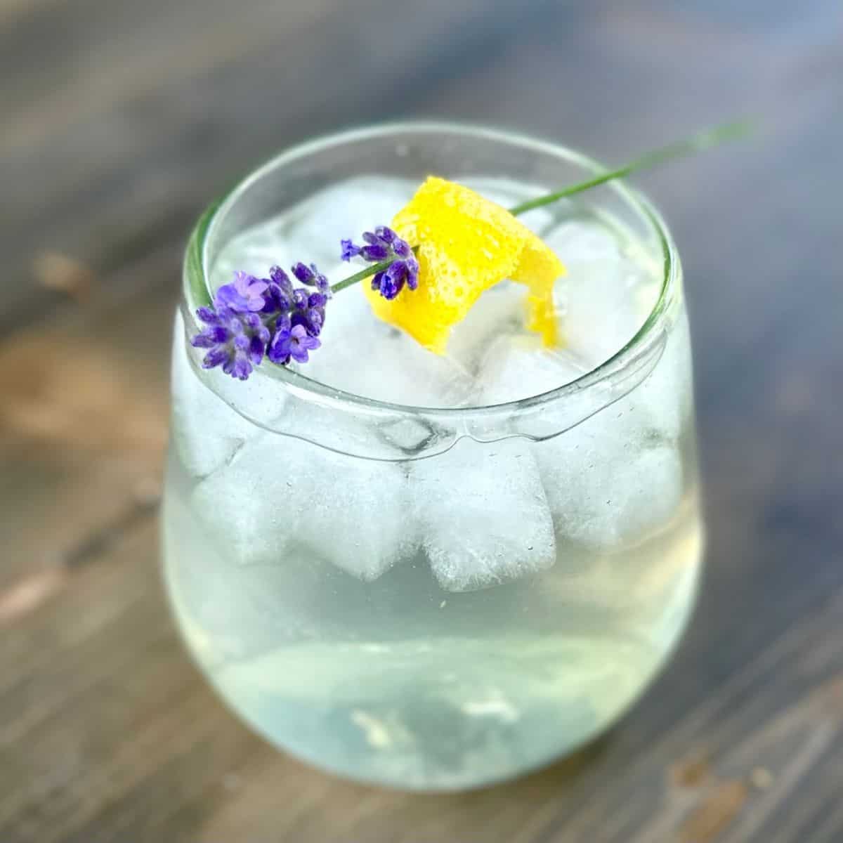 Glass filled with cocktail garnished with lavender and lemon rind. Sitting on a wooden surface.