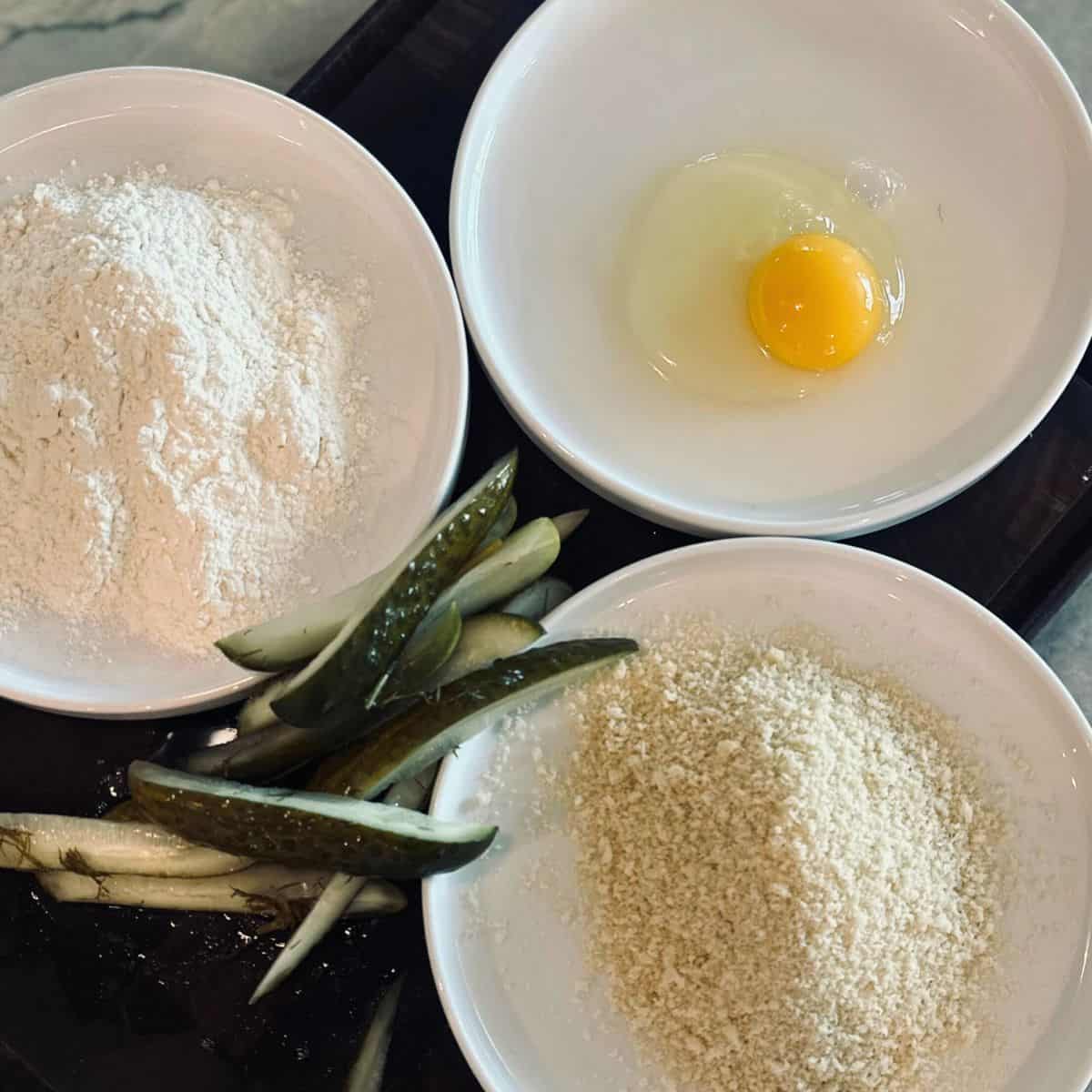 3 Plates with flour, egg and panko as well as sliced pickles.
