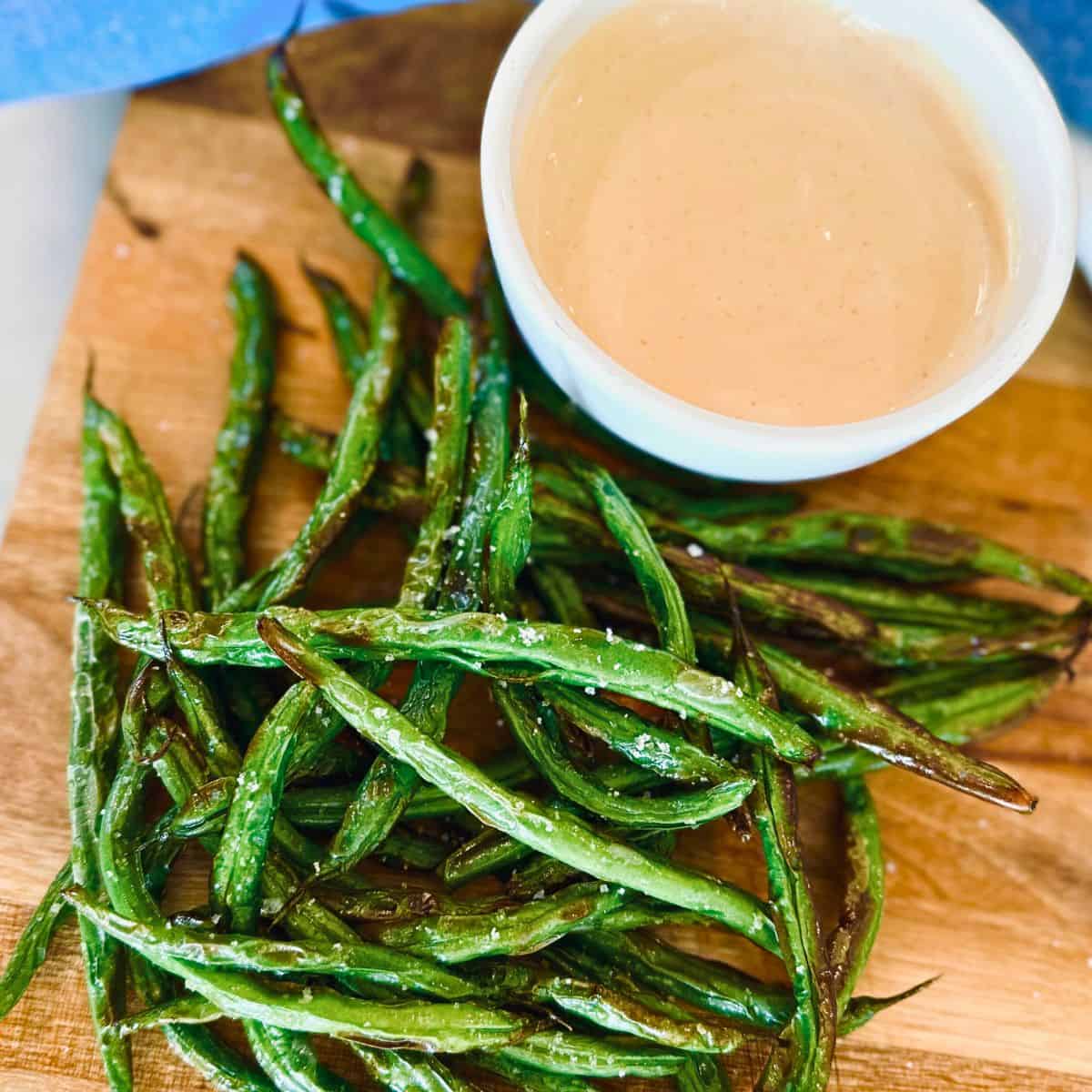 Air fryer green beans with dipping sauce on a wooden serving platter.